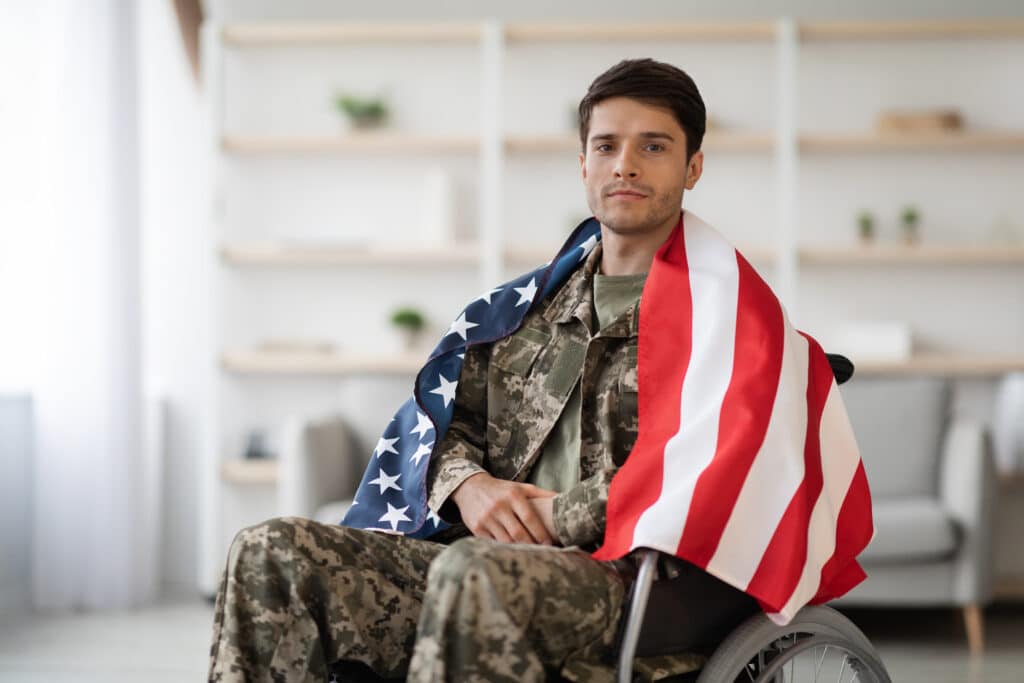 Young military guy wearing uniform sitting in wheelchair with flag of the US on his shoulders, home interior, panorama with copy space. Patriotism and military veterans concept